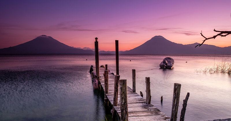 Wooden dock leading into Lake Atitlán at sunset with volcanoes in the background and a boat floating on calm water — serene Guatemala landscape.