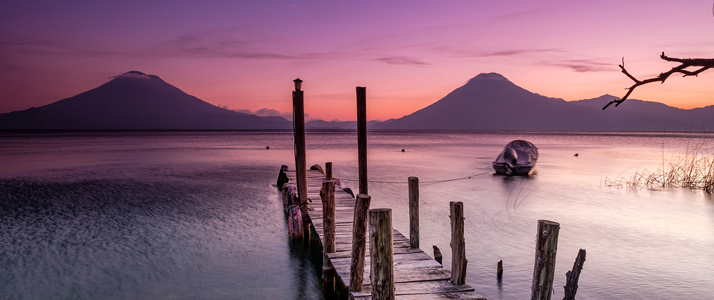 Wooden dock leading into Lake Atitlán at sunset with volcanoes in the background and a boat floating on calm water — serene Guatemala landscape.