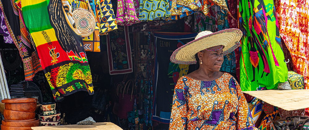 Woman in colorful traditional clothing and sun hat at an African market stall with vibrant textiles and handmade crafts — local culture and fashion.