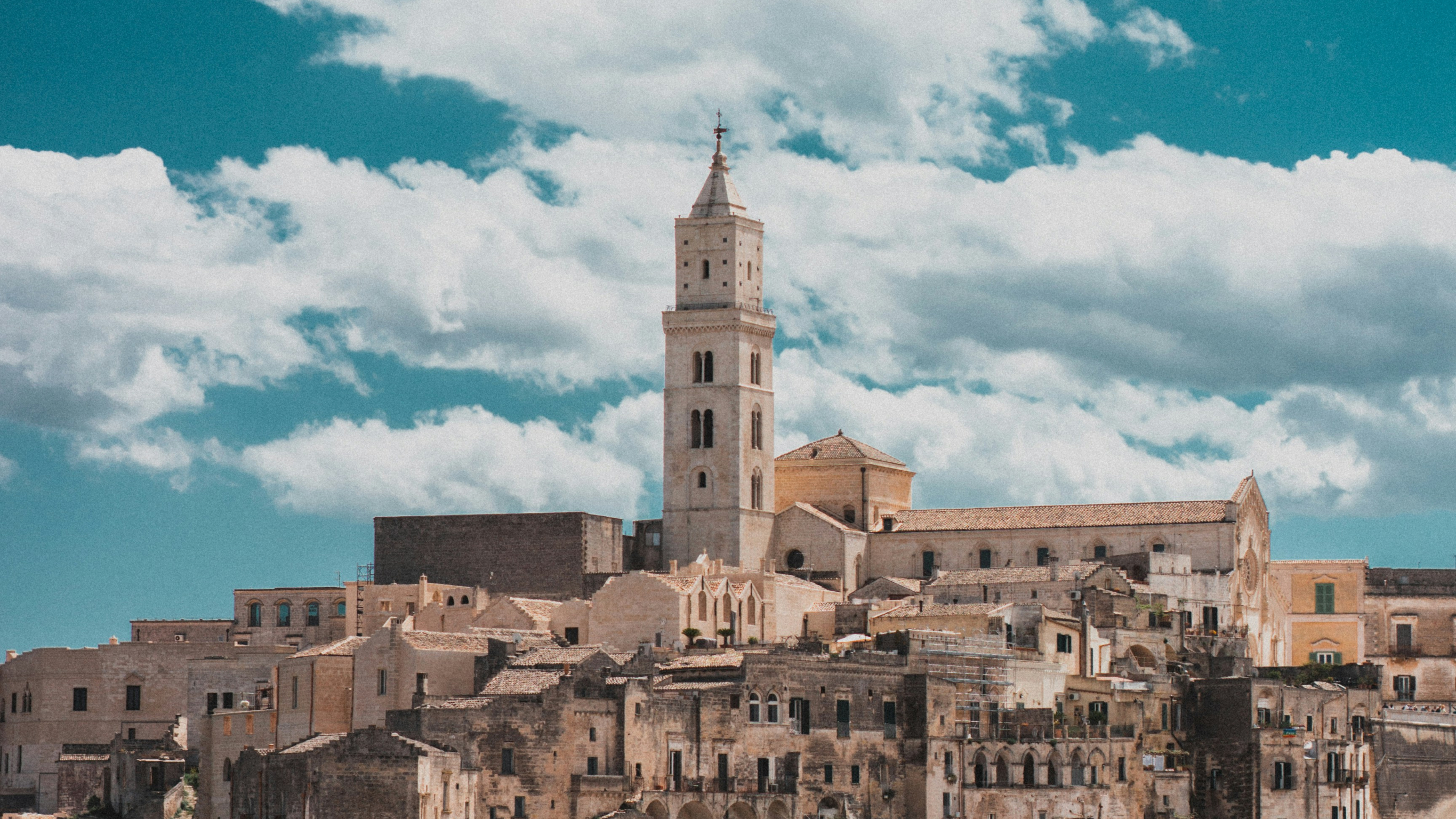 Historic stone buildings and tall bell tower of Matera, Italy under a bright blue sky with scattered clouds — UNESCO World Heritage hill town.