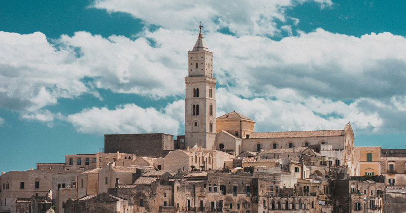 Historic stone buildings and tall bell tower of Matera, Italy under a bright blue sky with scattered clouds — UNESCO World Heritage hill town.