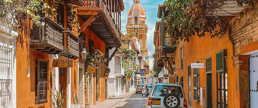 Colorful colonial street in Cartagena, Colombia with balconies, vibrant walls, and the iconic clock tower cathedral in the background — lively historic cityscape.