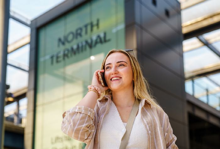 Smiling woman on the phone at Gatwick North Terminal calling Park & Deliver to have her car returned by Bubble Parking, with the terminal visible in the background