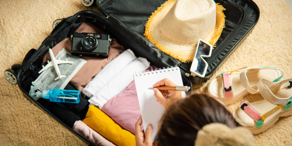 An open suitcase on a carpet containing neatly folded clothes, a straw hat, camera and model aeroplane, while someone writes in a notepad—illustrating cheap travel inurance.