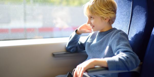 A young child with blonde hair sitting by a train window, smiling as they gaze out at the passing landscape bathed in sunlight.