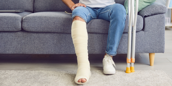A person seated on a grey sofa wearing jeans and a full-leg plaster cast, with a pair of crutches propped upright against the sofa beside them.