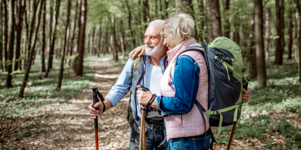 An elderly couple in their 70s with backpacks and walking poles embracing on a leafy forest trail, illustrating travel insurance for over 70’s.