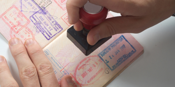A hand stamping a passport page filled with colourful visa and entry stamps, while another hand holds the passport steady against a white background.