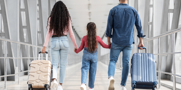 Back view of a family walking through an airport terminal. The parents, each pulling a suitcase, hold hands with their young daughter in the middle. They are casually dressed in denim outfits, and the setting appears to be a modern, well-lit hallway with large windows