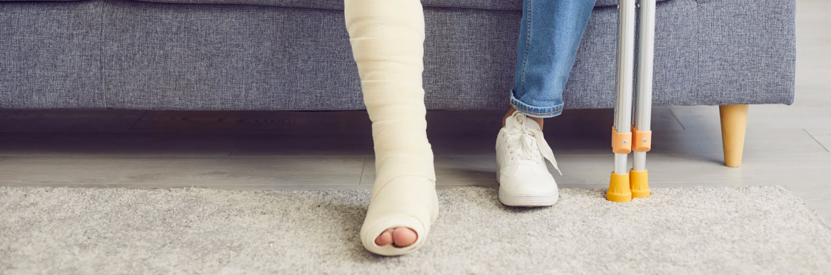 A person with their leg in a plaster cast, sitting on a sofa with crutches beside them – a visual representing the need for travel insurance for broken bones.