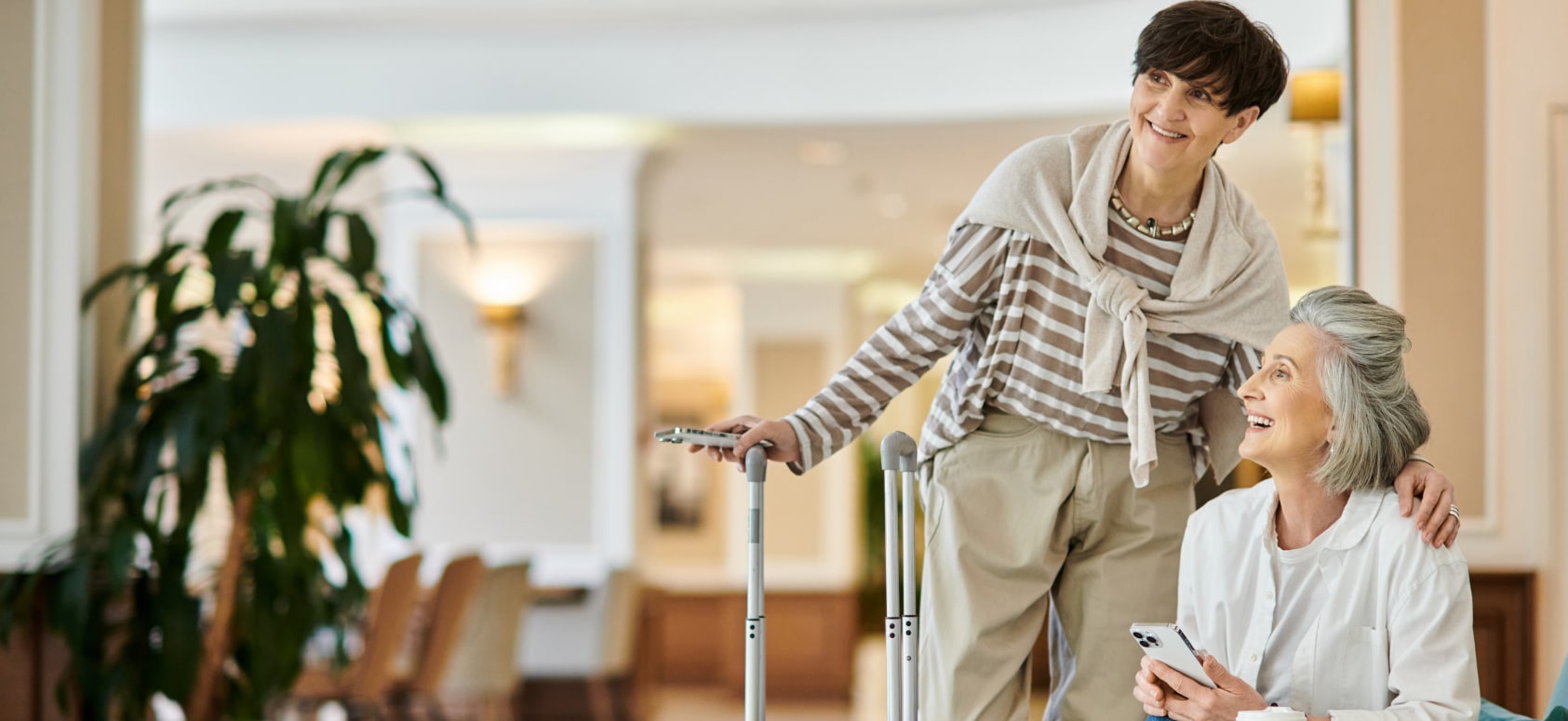 Two smiling older women with luggage in a bright hotel lobby, one sitting and holding a phone while the other stands beside her with a suitcase.