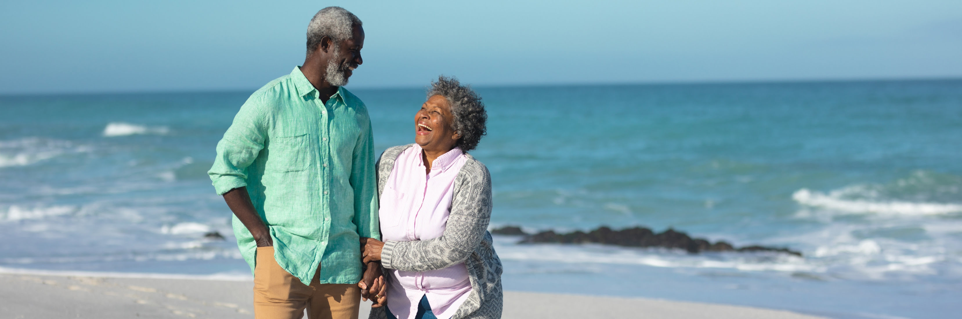 Smiling senior couple walking hand in hand along the beach with the ocean in the background.