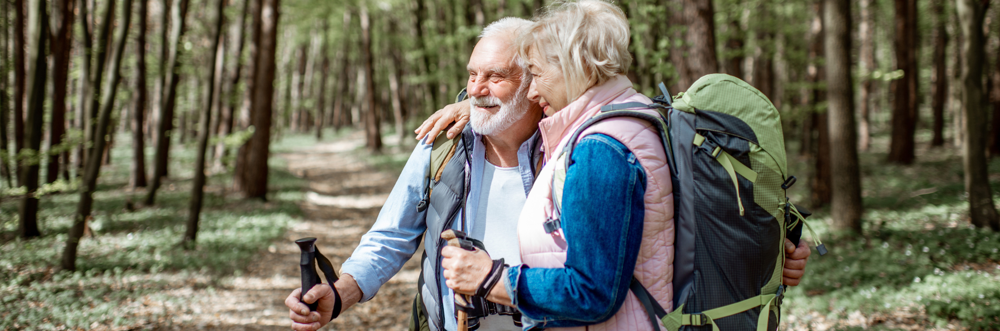 Smiling couple in their 80s hiking through a sunlit forest with backpacks and walking sticks, illustrating the security offered by travel insurance for over 80's