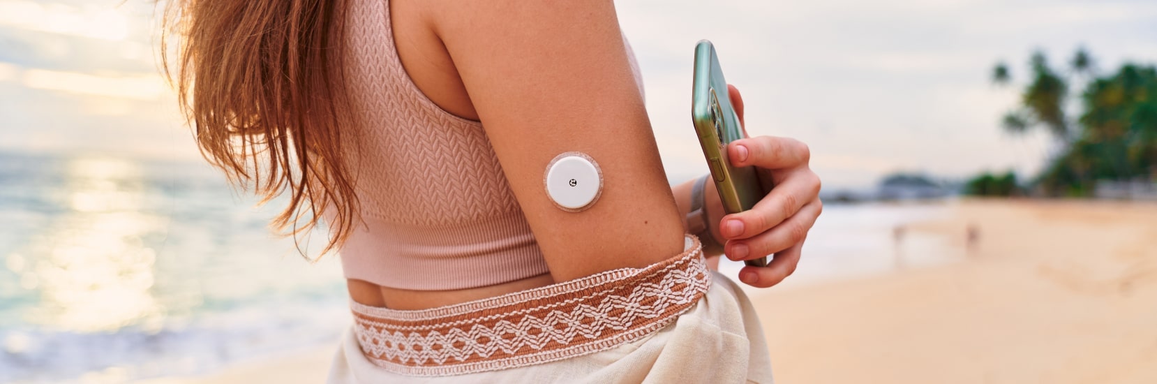 Person on a beach holding a phone with a visible glucose monitoring device on their arm..
