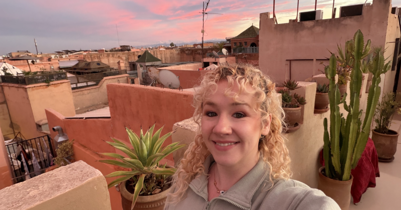 Smiling solo female traveller taking a selfie on a rooftop terrace at sunset, with potted plants and warm pink sky over a city skyline — relaxed holiday vibe.