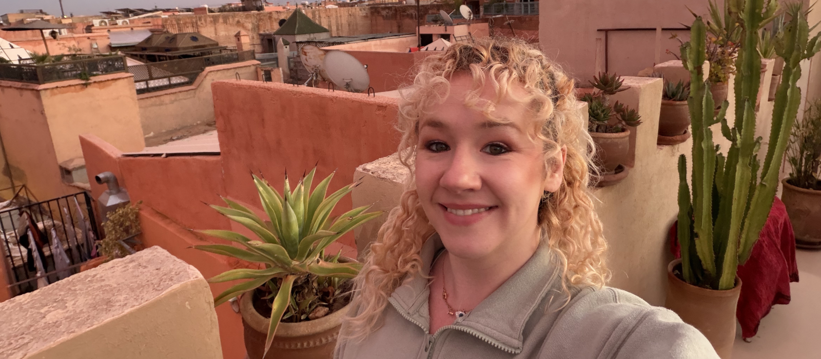 SSmiling solo female traveller taking a selfie on a rooftop terrace at sunset, with potted plants and warm pink sky over a city skyline — relaxed holiday vibe.