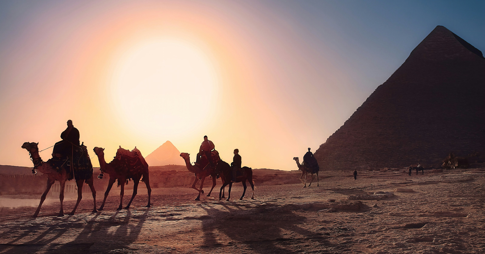 Silhouettes of camel riders near the Pyramids of Giza at sunset, with golden desert light and ancient Egyptian landmarks.