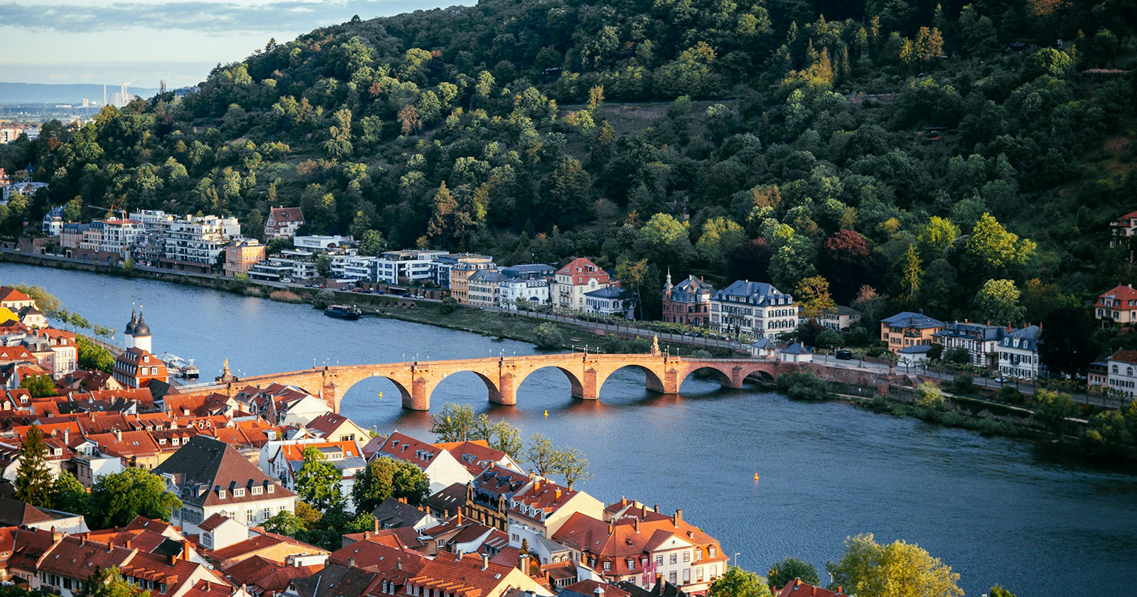 Scenic view of Heidelberg’s Old Bridge crossing the Neckar River, with red-roofed buildings and forested hills in Germany.