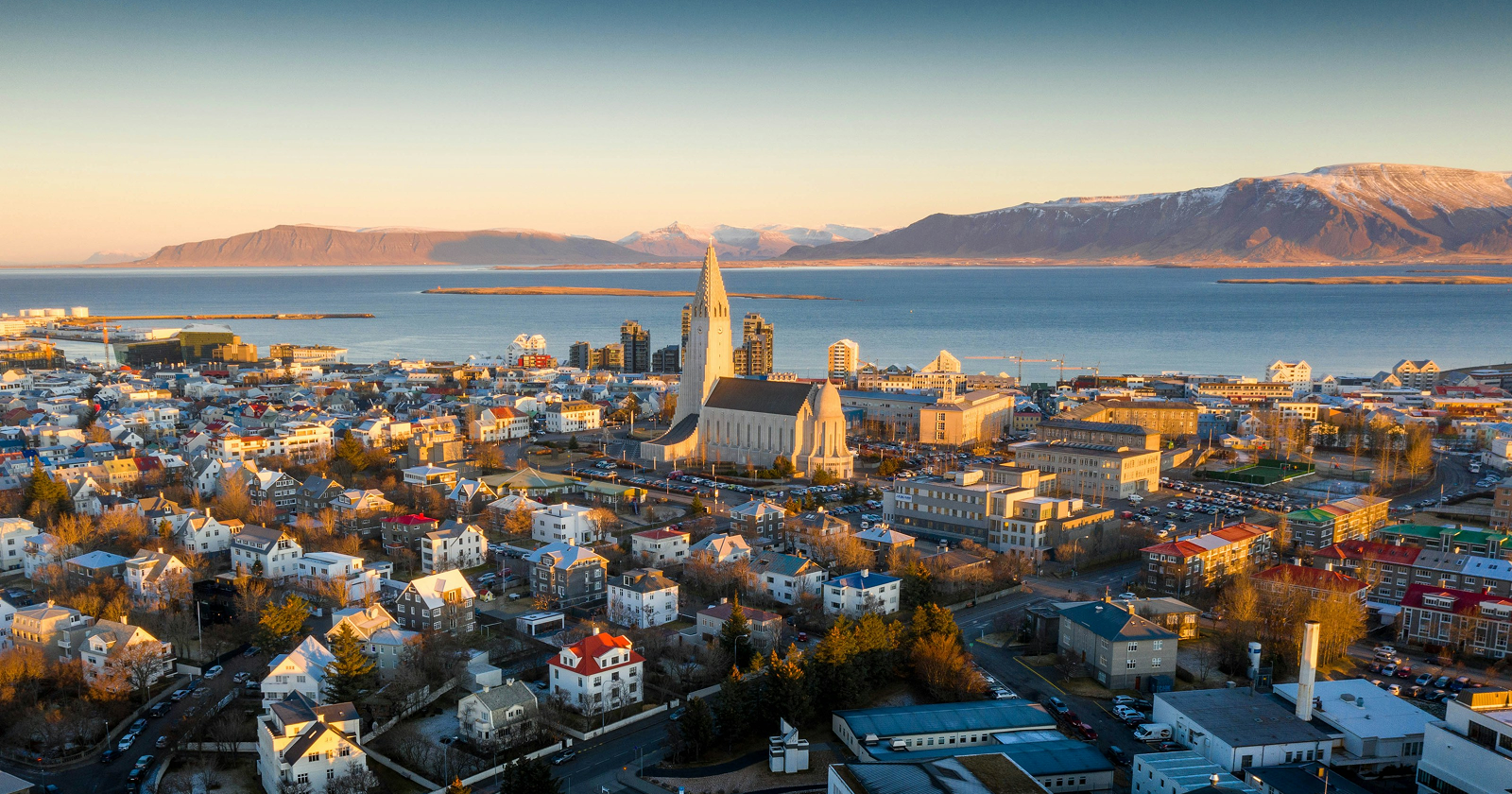 Aerial view of Reykjavik, Iceland, with Hallgrímskirkja church, colorful homes, coastal waters, and snow-capped mountains at sunset.