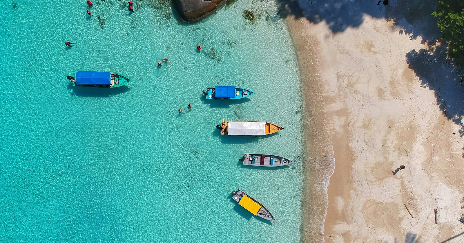 Aerial view of a tropical beach with turquoise water, small boats, swimmers, and white sand shoreline — relaxing island vacation scene.