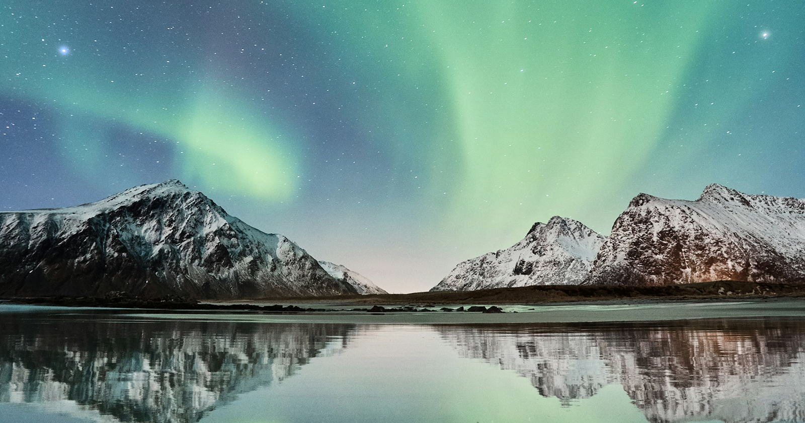 Green northern lights above snowy mountains reflected in a calm lake under a starry Arctic night sky. 