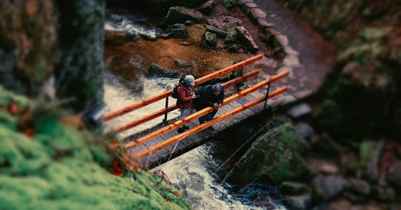 Two hikers standing on a wooden bridge above a rushing forest stream surrounded by mossy rocks and greenery.