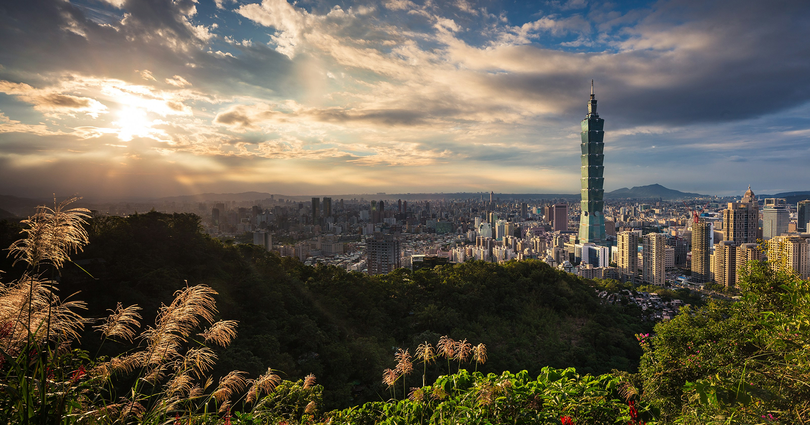 Taipei skyline at sunset with Taipei 101 tower, dramatic clouds, and lush green hills overlooking the modern cityscape.