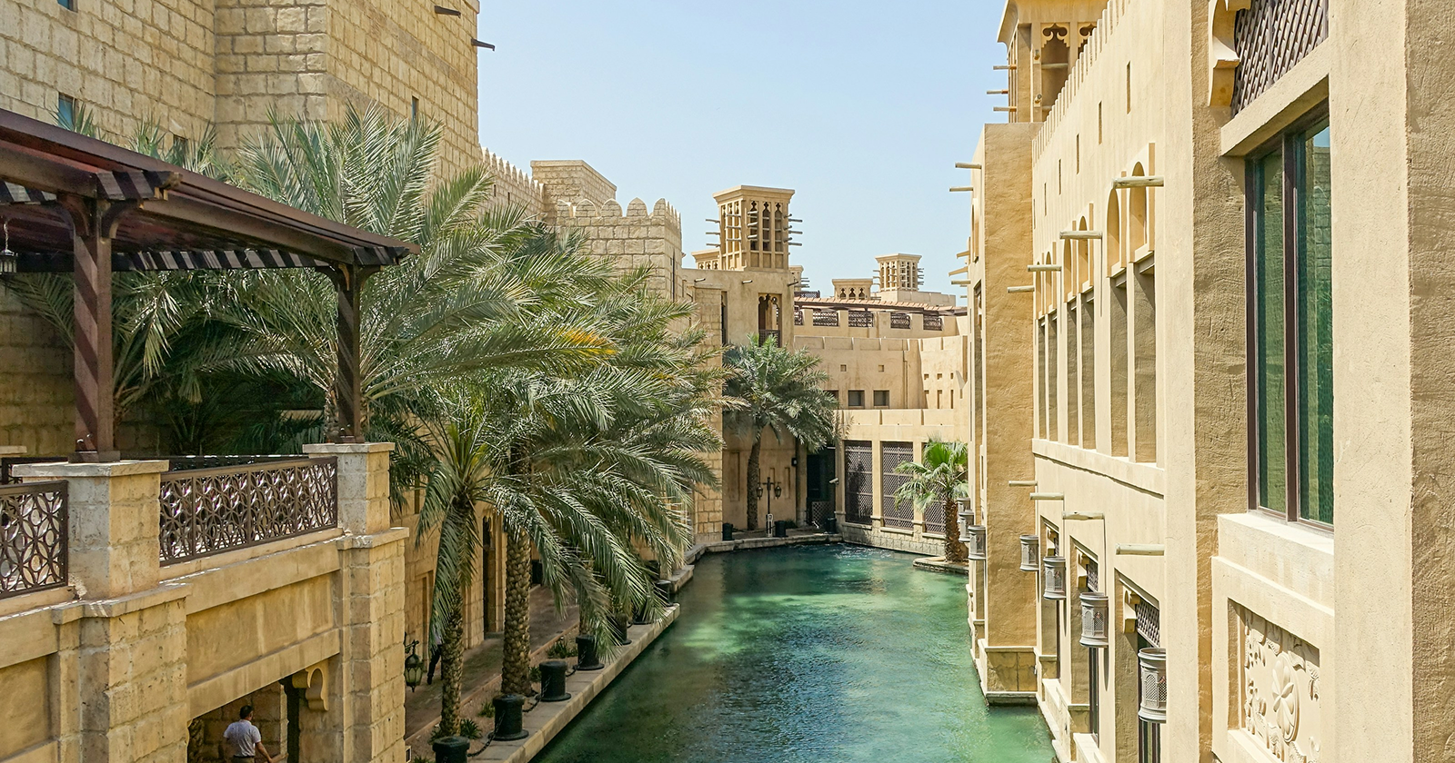 Traditional Arabian-style buildings with palm trees and turquoise canal water in a luxury Middle Eastern resort setting.