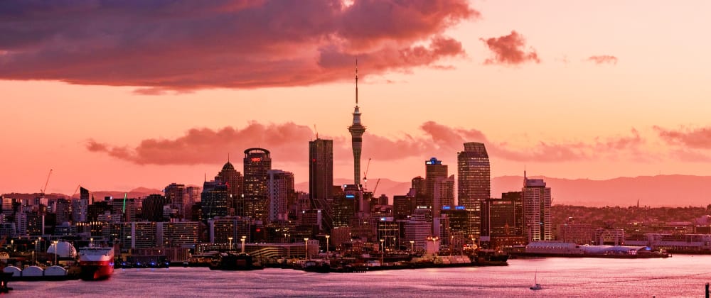 Auckland skyline at sunset with the Sky Tower above waterfront buildings and harbour, beneath dramatic pink and orange clouds.