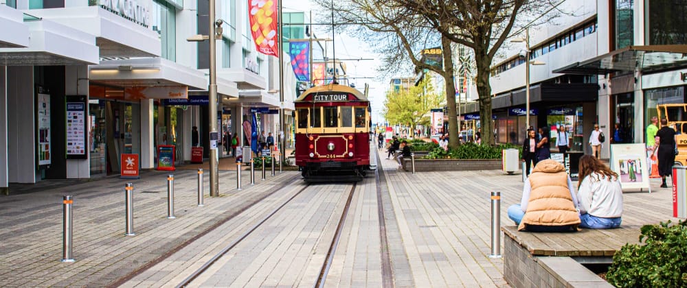 Red vintage city tour tram traveling along a pedestrian shopping street, with shops, trees, and people in the city centre.