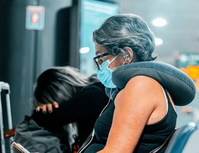 Frau mit Brille und Maske sitzt mit Nackenkissen auf den Schultern am Flughafen und liest
