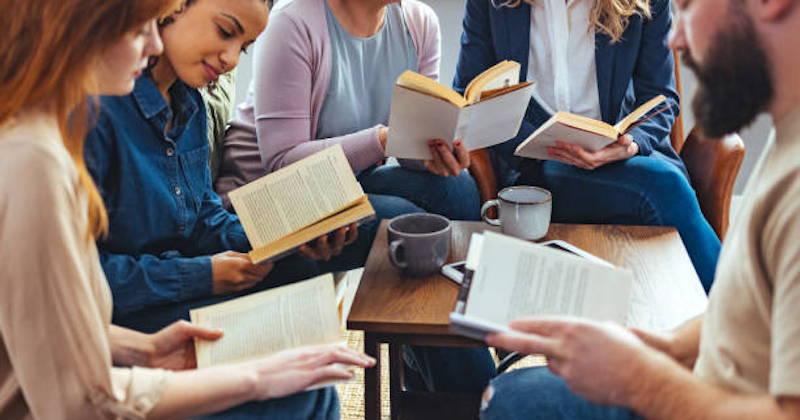 Diverse group of people sitting in a cosy setting, reading books around a coffee table with mugs – casual book club meeting.